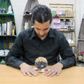 Un homme concentré devant une Boule de Cristal Sur Support - 100 mm, symbole fort du monde ésotérique. Cet accessoire spirituel sert aussi bien pour des rituels sur un autel ésotérique que pour une décoration mystique. La boule cristal 100 mm est parfaite comme objet de divination ou élément décoratif dans une ambiance spirituelle.