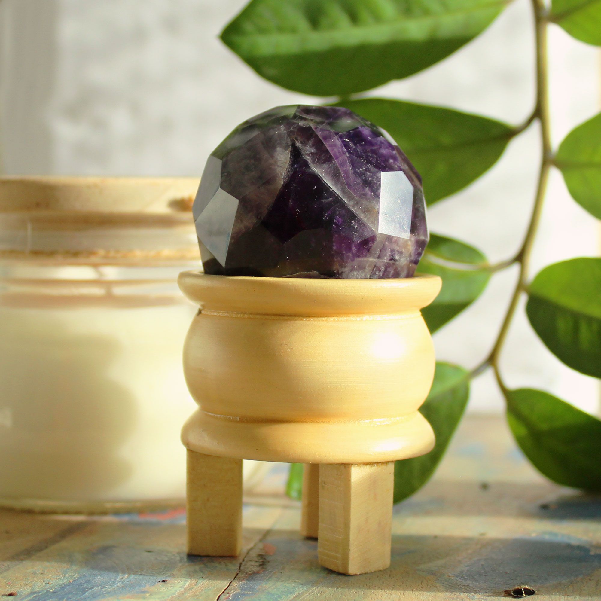 Boule De Guérison En Améthyste Facetée sur un support en bois, entourée de verdure, cette sphère améthyste en pierre naturelle est idéale pour la méditation et la lithothérapie. Sa beauté naturelle favorise la sérénité et l'intuition, tout en activant le chakra couronne et le troisième œil. Une magnifique pièce de décoration spirituelle de la collection Aemilia Mystica.