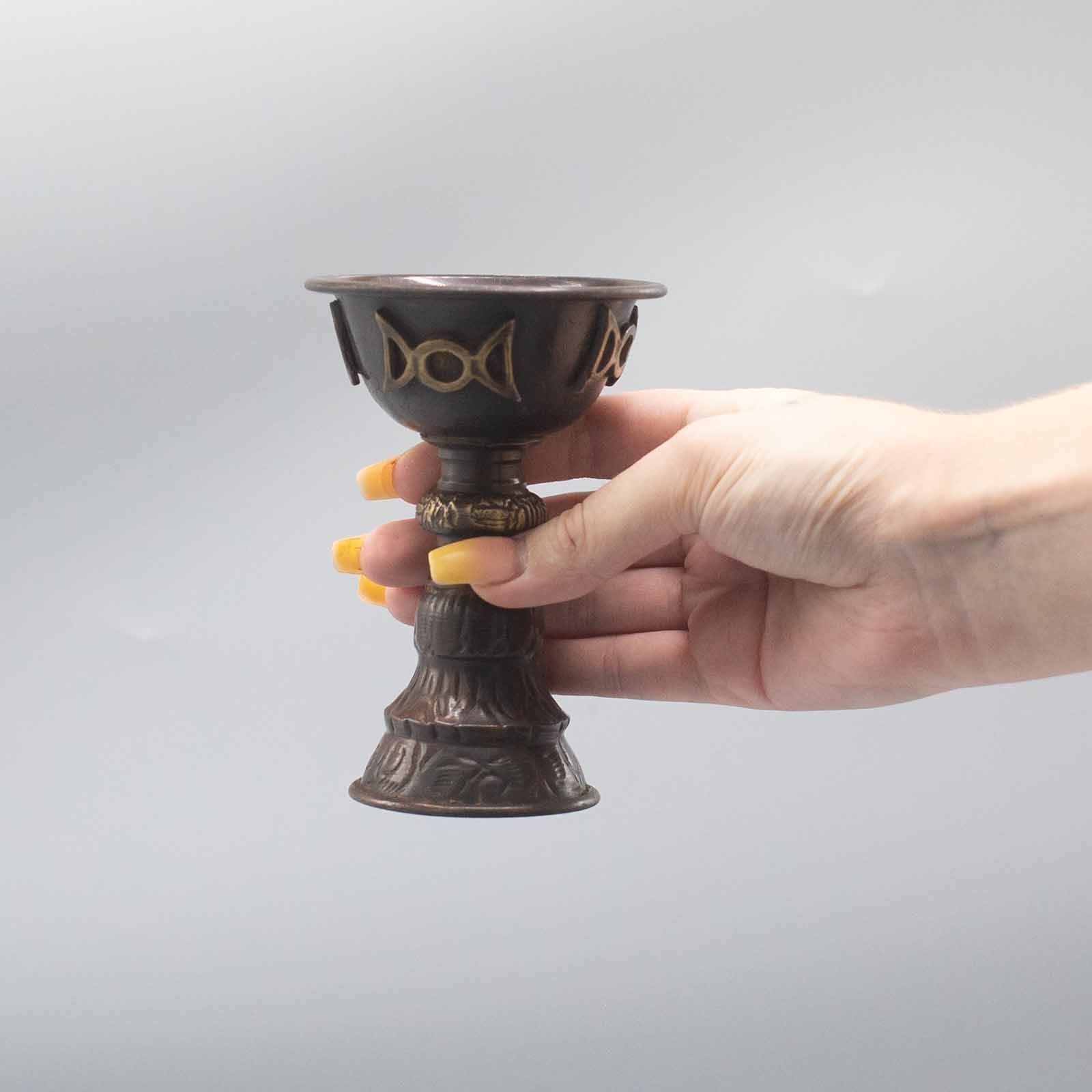Calice Rituel en Cuivre Antique Triple Lune, tenu à la main, illustrant sa taille parfaite pour un usage personnel. Ce gobelet rituel est une pièce essentielle pour vos cérémonies spirituelles, vous accompagnant dans vos offrandes. Parfait comme accessoire rituel, il incarne le féminin sacré et la magie moderne à travers son design en cuivre antique.