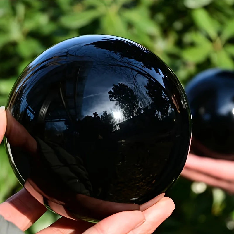Boule en obsidienne noire naturelle polie tenue dans la main, avec autres sphères en pierre en arrière-plan 12 cm